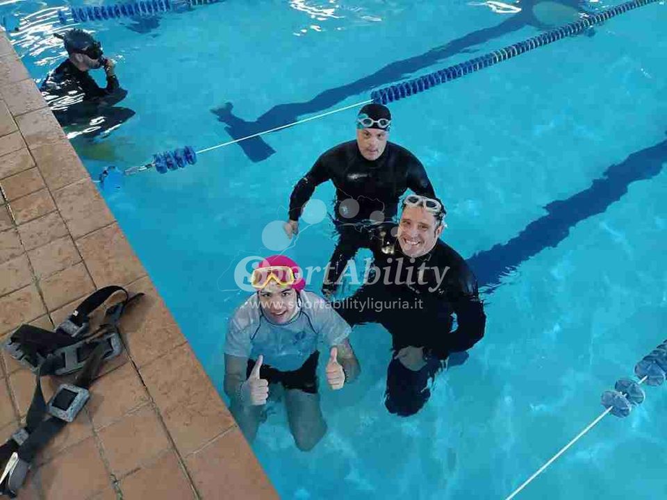 foto ripresa dall'alto del bordo piscina, si vedono in acqua un ragazzo che alza i pollici con maglietta del Centro e cuffietta lilla, un istruttore sorridente dietro di lui e un altro ragazzo sorridente che indossa una muta dietro di loro