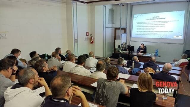 Un'aula piena di partecipanti durante una lezione frontale. Una persona seduta alla cattedra sta spiegando con l'ausilio di una presentazione proiettata su uno schermo, relativa al Centro Iperbarico di San Martino. Sulla proiezione si vedono vari punti elencati, mentre il logo di Genova Today è visibile in basso a destra.