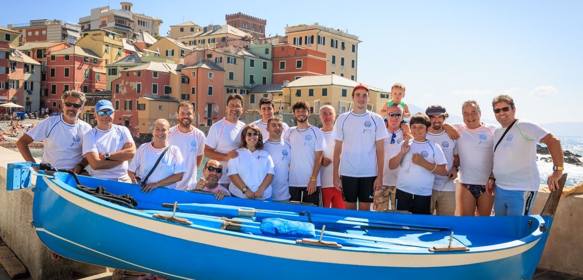 Allievi e istruttori del Centro in posa vicino a una barca azzurra, indossando magliette bianche con il logo del Centro Subacqueo. Sullo sfondo si vedono edifici colorati in stile mediterraneo e il mare. Tra i partecipanti ci sono uomini, donne e bambini sorridenti.